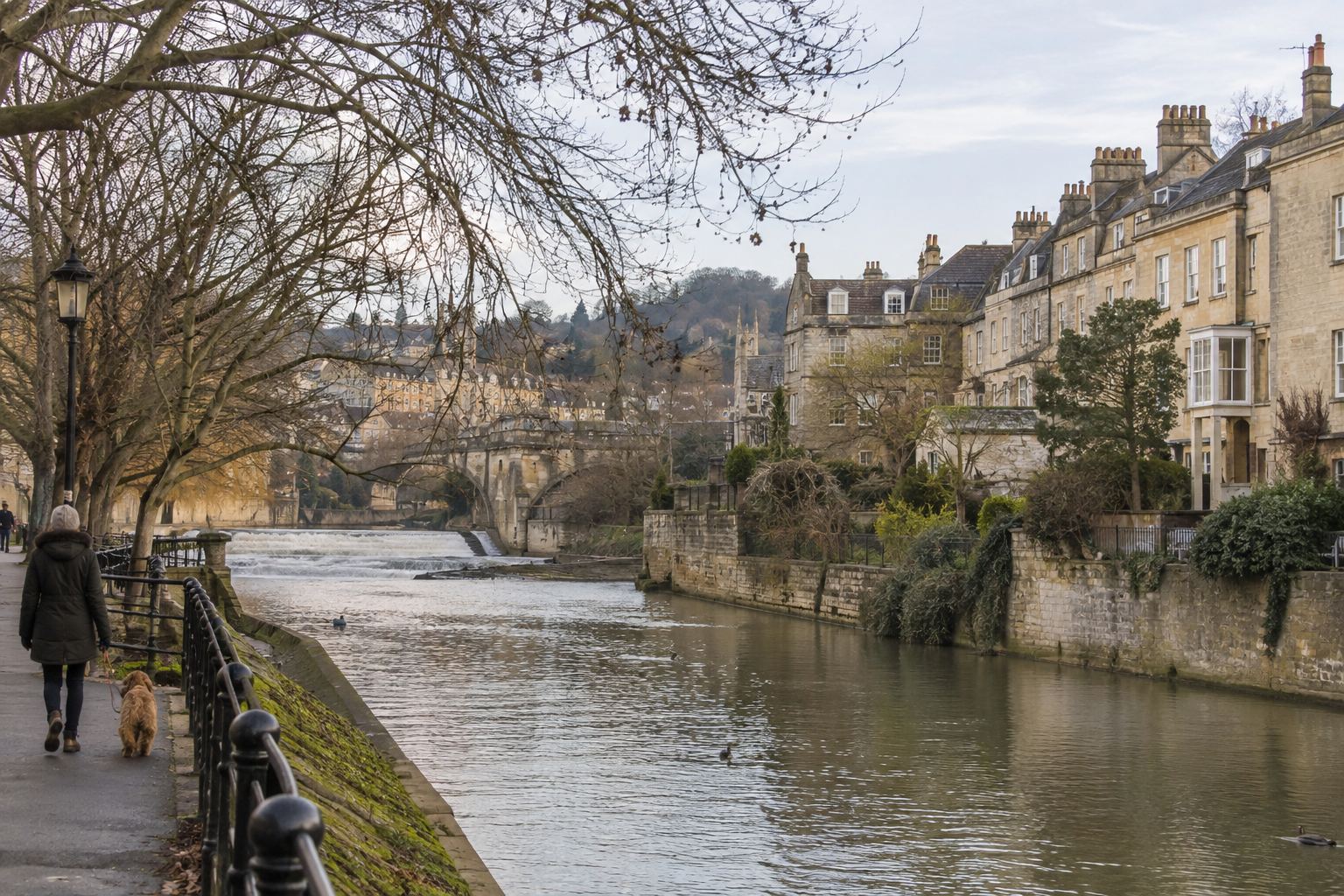 Stone townhouses along a riverside path with a woman walking a dog